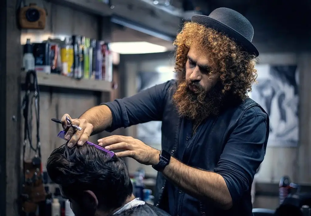 Een man met een baard en een hoed knipt het haar van een andere man in Enschede